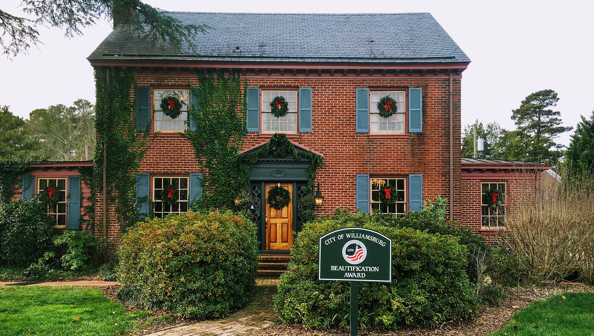 A brick house adorned with festive wreaths and surrounded by greenery, featuring a sign for a beautification award.