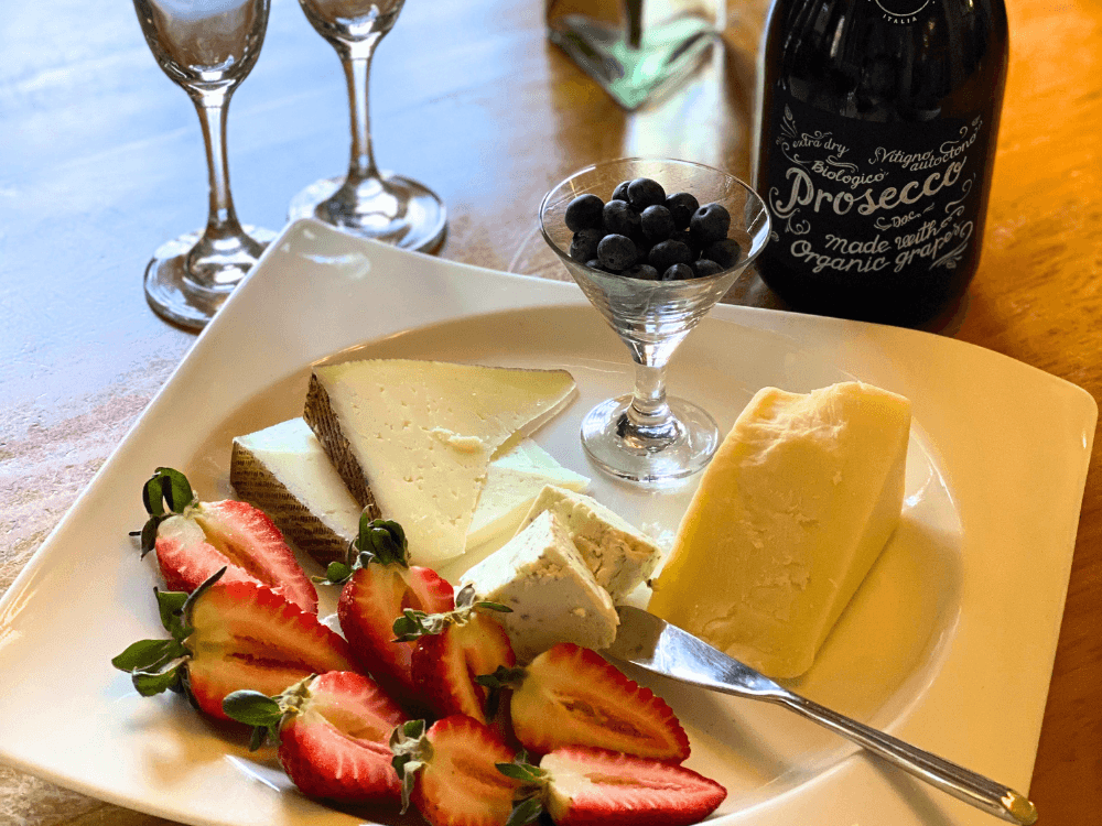 A plate of assorted cheeses, fresh strawberries, and blueberries paired with a bottle of Prosecco.