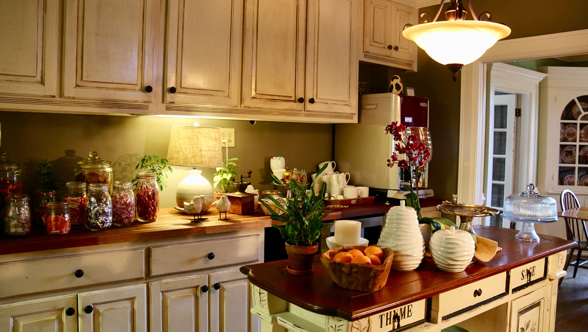 A cozy kitchen featuring wooden cabinets, jars of colorful ingredients, fresh fruits, and decorative plants on the countertop.